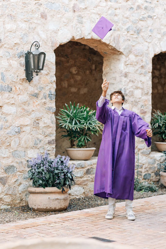 Notre Dame Prep High School Senior in Scottsdale, AZ standing in the courtyard at Silverleaf Country Club wearing a purple graduation gown and tossing his graduation cap in the air. 