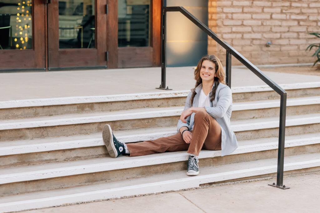 Cave Creek high School senior in brown pants, white t-shirt, and gray button up shirt sitting on concrete steps smiling at the camera. 