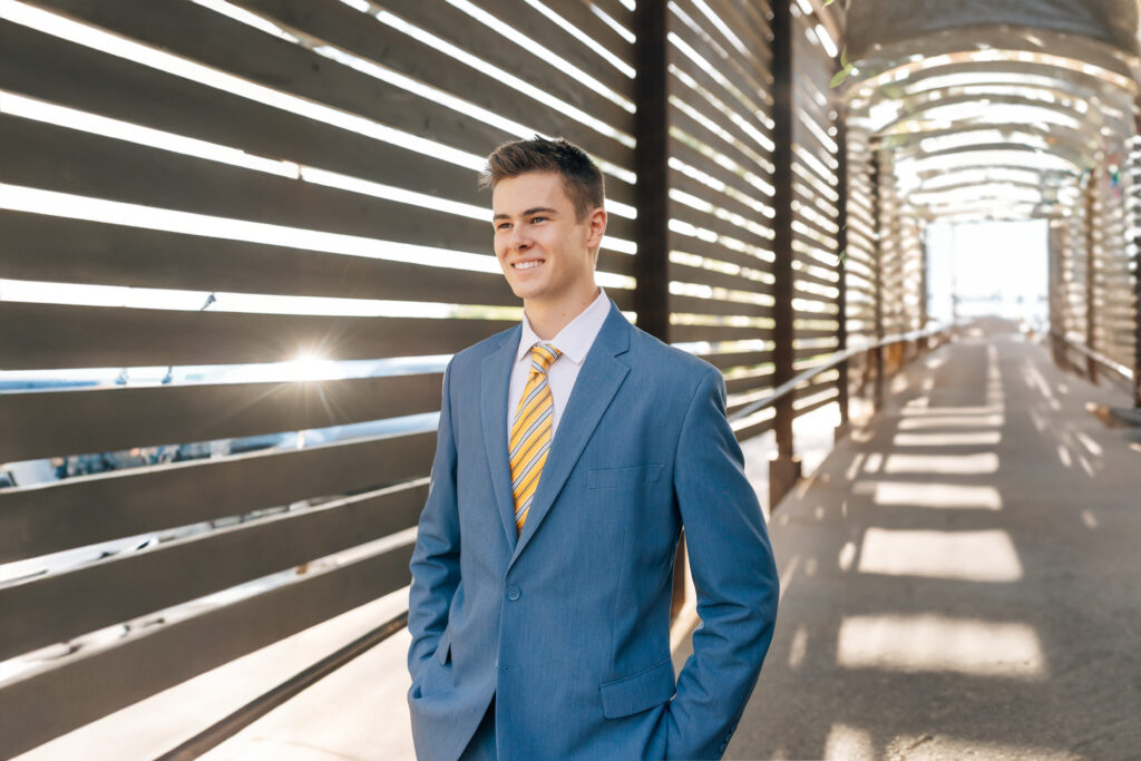 Mountain View Mesa, AZ high school senior guy wearing a blue suit, white dress shirt, and yellow striped tie while walking down a tunnel. The sun is to the left shining through a slat in the tunnel as he looks off ahead with a  smile. 