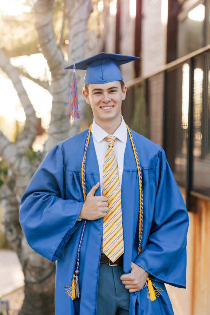 High school senior boy smiling at the camera and wearing a white collared shirt, yellow and blue striped tie, blue dress pants with a brown belt, and his blue cap and gown with a gold honor cord over his shoulders. 