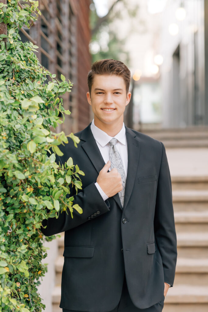 Mountain View Mesa, AZ high school senior guy wearing a dark gray suit, white dress shirt, and light gray floral tie while standing near greenery in a suburban location. 