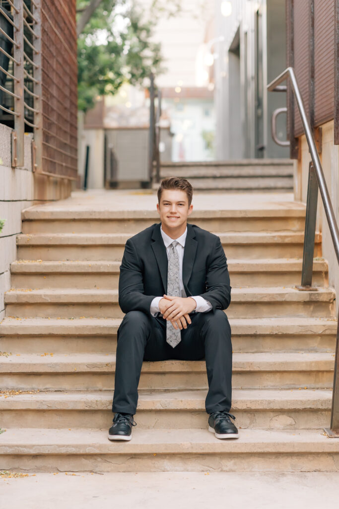 Mountain View Mesa, AZ high school senior guy wearing a dark gray suit, white dress shirt, and light gray floral tie while sitting on concrete steps in a suburban location. 