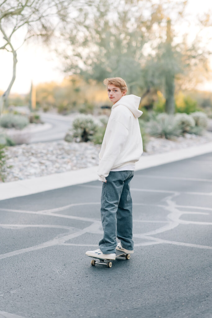 High school senior skater boy wearing gray corduroy pants and a white oversized hoodie as he looks over his shoulder toward the camera while skating away. 