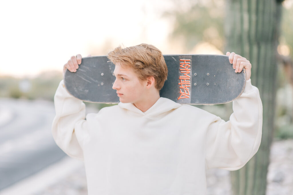 High school senior in Scottsdale, AZ holding his skateboard over his shoulders while looking to the left in a white oversized hoodie. 