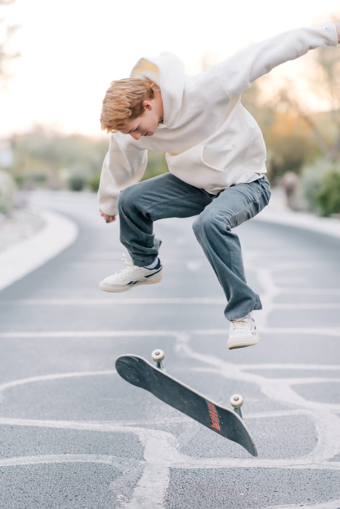 High school senior in Scottsdale, AZ wearing gray courduroy pants and a white hoodie doing a skate board trick. 