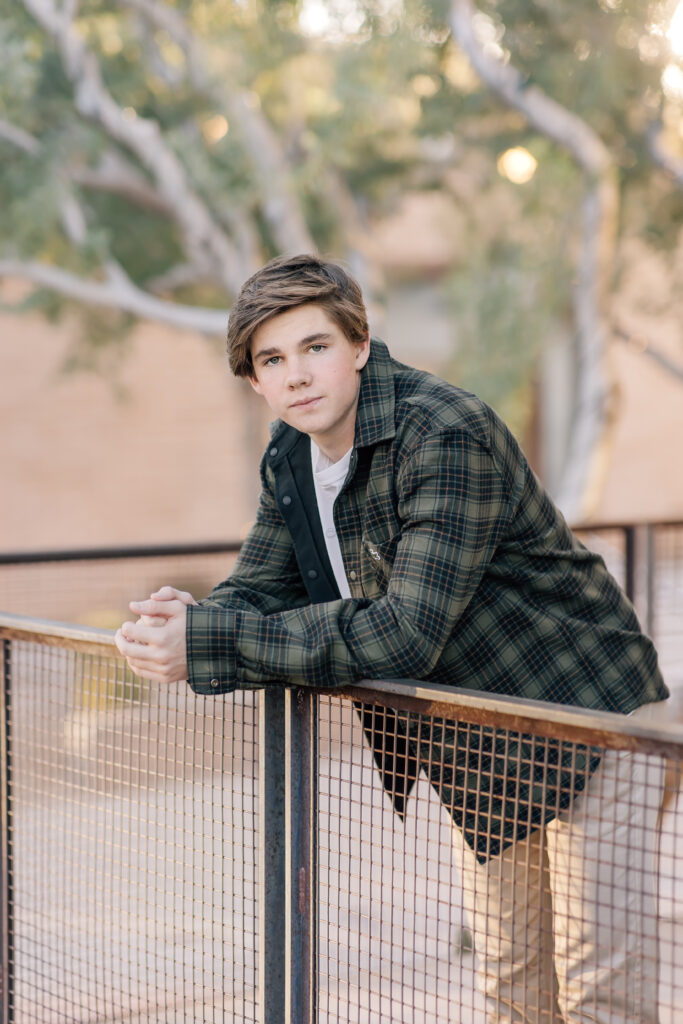 Scottsdale high School senior in khaki pants, white t-shirt, and olive jacket leaning over a balcony wall and looking at the camera. 