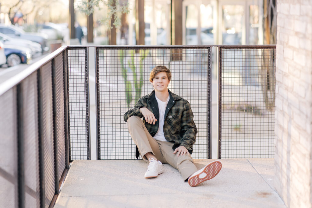 Scottsdale high School senior in khaki pants, white t-shirt, and olive jacket sitting while leaning against a balcony wall. 