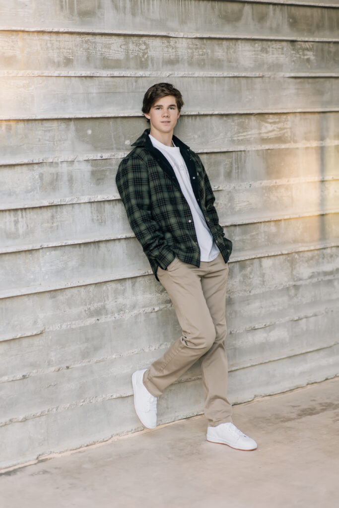 Scottsdale high School senior in khaki pants, white t-shirt, and olive jacket leaning against a concrete wall. 