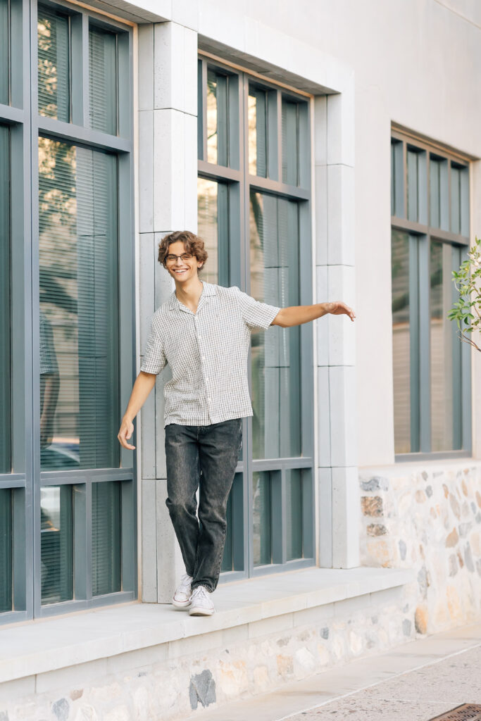 Chaparral High School Scottsdale, AZ high school senior in black denim pants and a plaid gray and white button up shirt with glasses is walking along a concrete wall playfully smiling at the camera. 