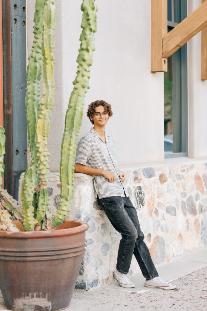 Chaparral High School Scottsdale, AZ high school senior in black denim pants and a plaid gray and white button up shirt with glasses is leaning against a concrete wall. 