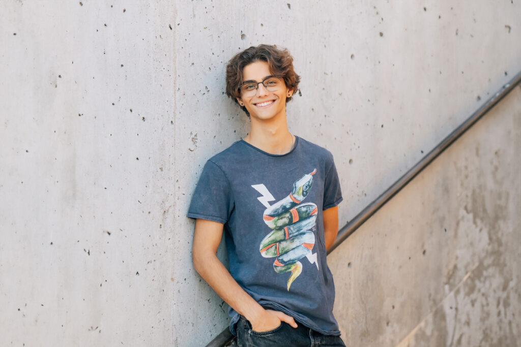 Chaparral High School Scottsdale, AZ high school senior in black denim pants and a blue graphic t-shirt with glasses is leaning against a concrete wall. 