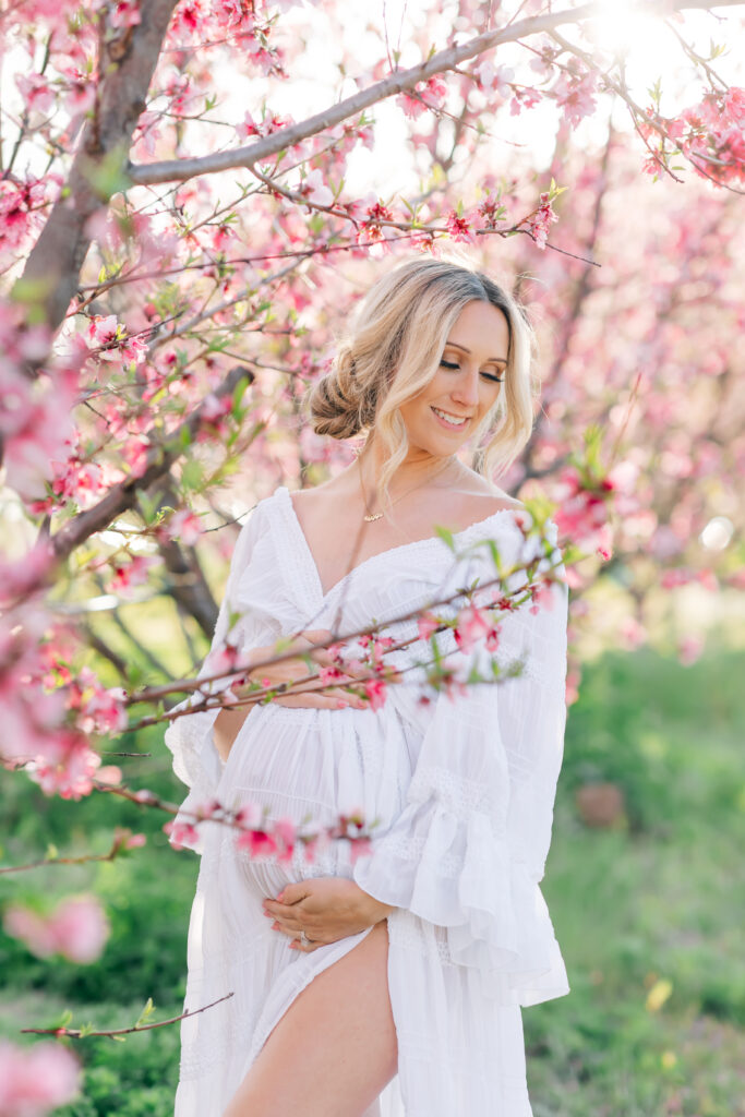 Expecting mother in a white dress cradling her baby bump in a peach blossom orchard, glowing in soft light — a serene moment from Nest: A Luxury Maternity & Newborn Workshop.