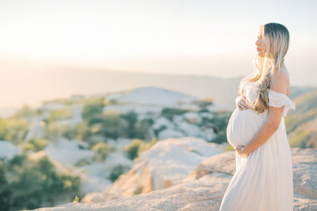 Pregnant woman in a flowing white dress, cradling her belly as she gazes over the horizon, bathed in soft golden light — capturing the warmth and serenity of motherhood with luxury maternity photography.